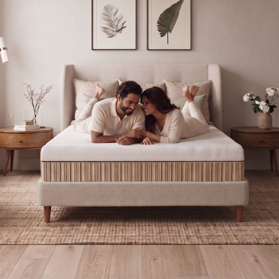 A couple lies on a bed with a Essentia stratami mattress and a beige upholstered frame in a softly lit bedroom. The scene includes wooden bedside tables, framed botanical wall art, and a woven rug.
