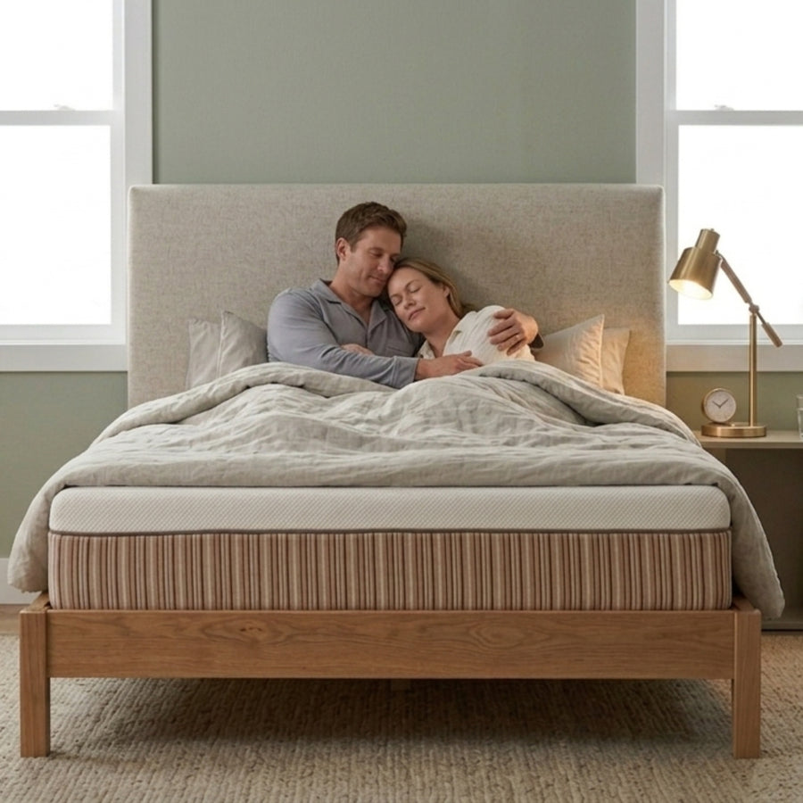A couple relaxes together on a wooden bed with a beige upholstered headboard, covered under a light gray quilted comforter in a softly lit bedroom.