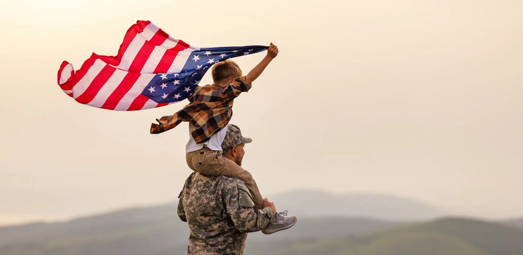 A man in a military uniform carrying a young boy on his shoulders who is holding up an American flag