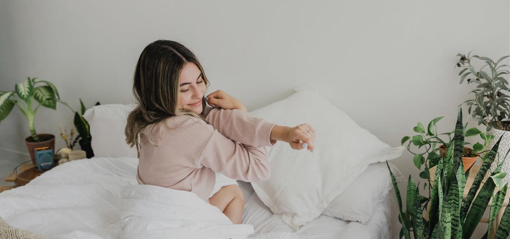 Woman wearing a pink sweater is stretching after waking up. Her bedroom is clean with a white duvet and green plants on both sides of the bed