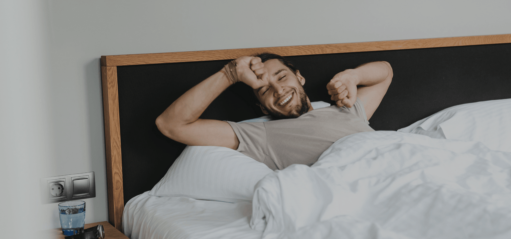 Man waking up and stretching while smiling in a bed with a dark headboard and fluffy white comforter