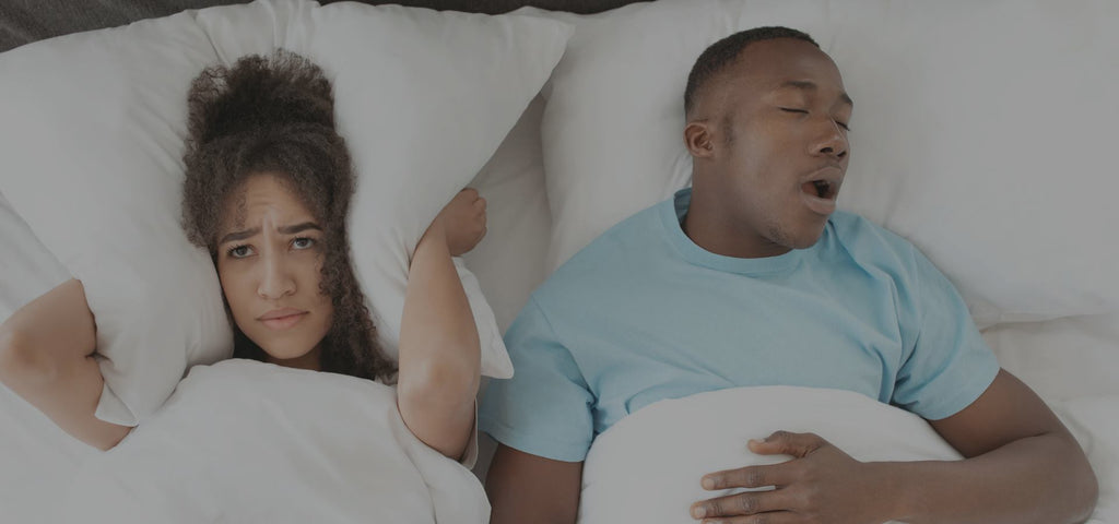 woman up and miserable holding her pillow to her ears, while her husband sleeps and snores loudly with his mouth open