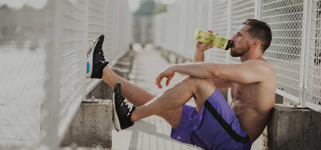 Man sitting down after a work out shirtless and drinking from a yellow bottle of water