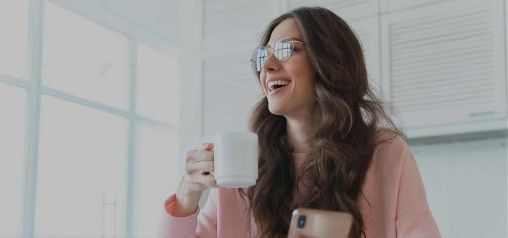 Beautiful woman with long hair smiling after waking up ready to take on the day. She has a cup of coffee and her cell phone in her hand.