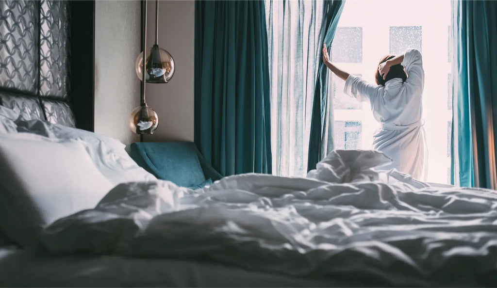 Woman waking up in a designer hotel stretching and drawing the curtains.