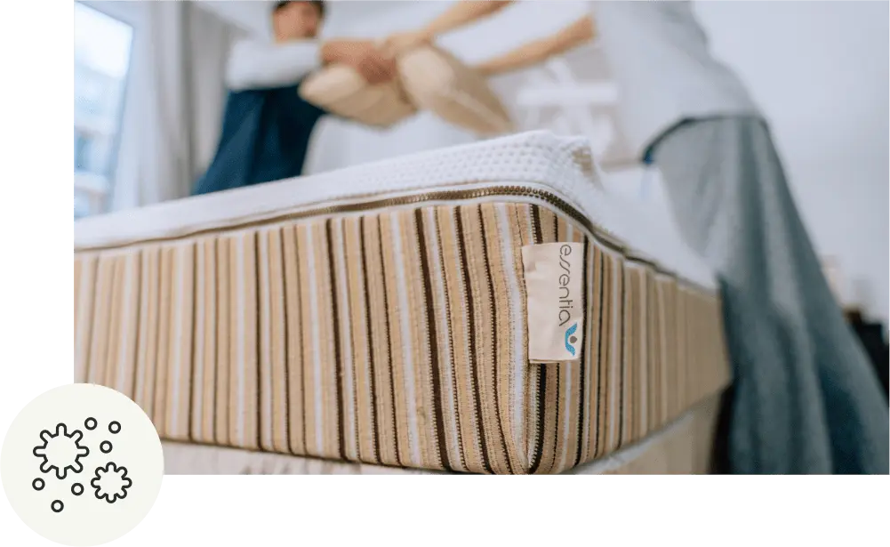 Close up view of the corner of an Essentia mattress. A couple helping each other to make the bed in the background.